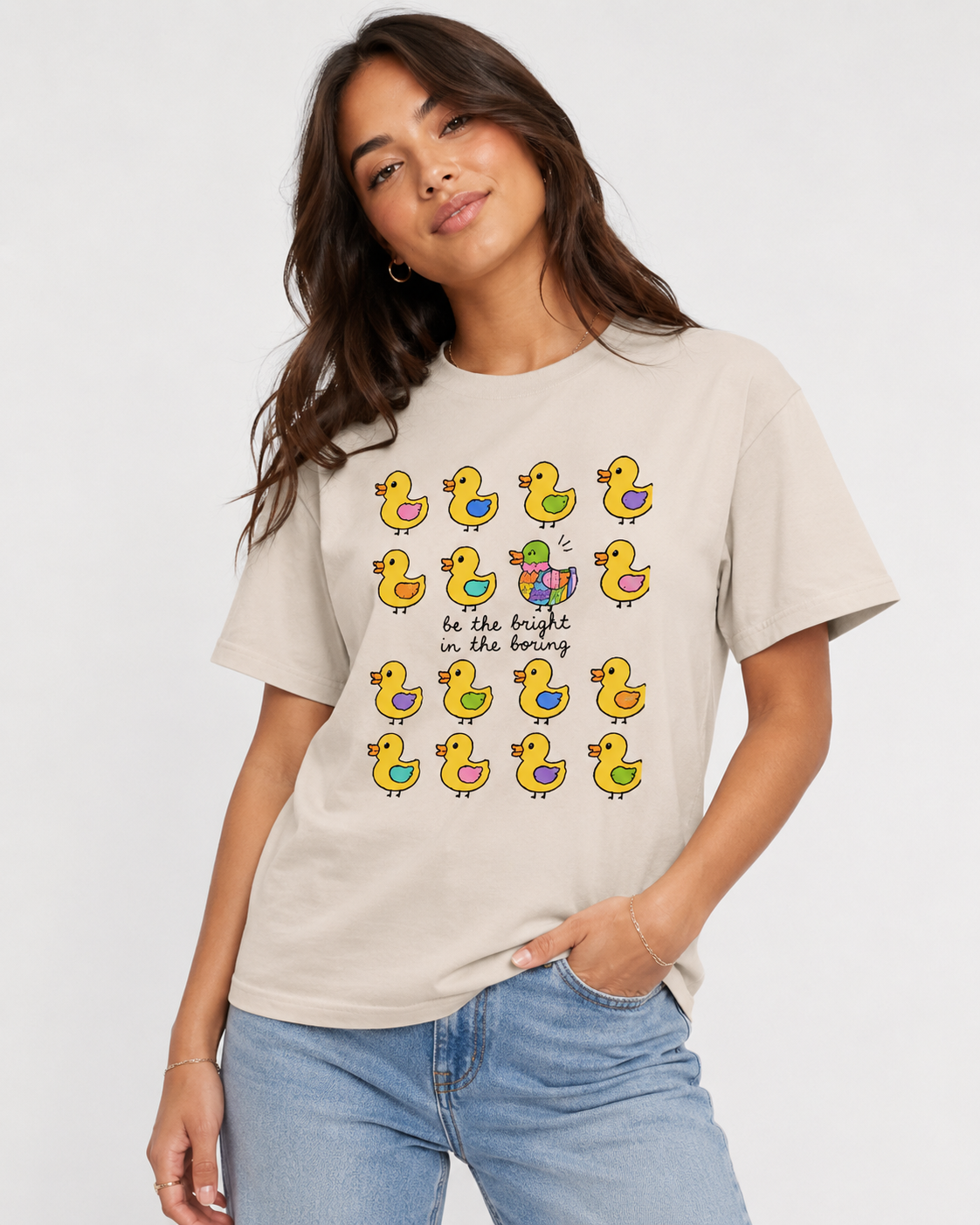 Woman wearing a beige t-shirt with colorful duck illustrations and text. text says be the bright in the boring 